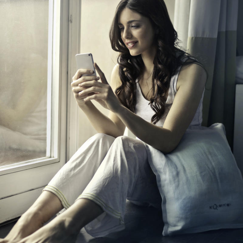 Junge Frau sitzt am Fenster und schaut lächelnd auf ihr Handy. Entspannte Atmosphäre.